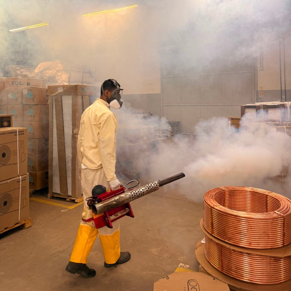 Técnico da DDTizaMax aplicando termonebulização em um galpão, de macacão, luvas e máscara de proteção